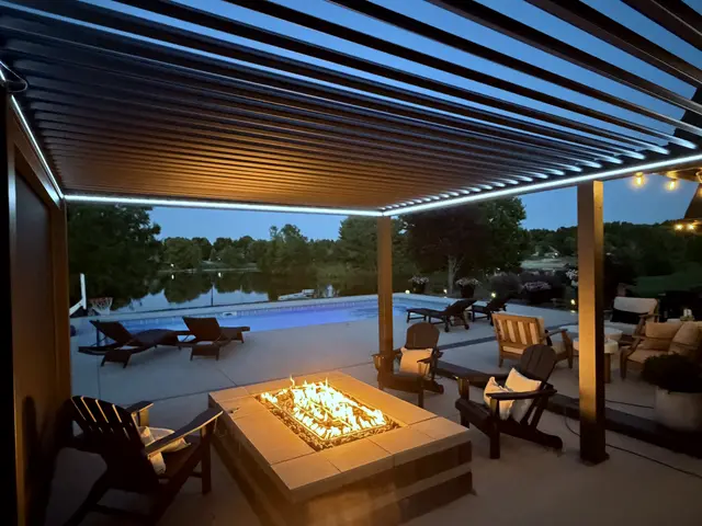 A finished louvered pergola with a fire pit and seating beside a pool, in the evening.